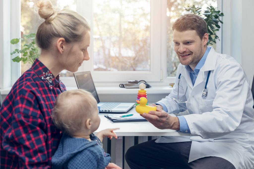 Bilde av en dame som sitter hos en lege med barnet sitt på fanget.